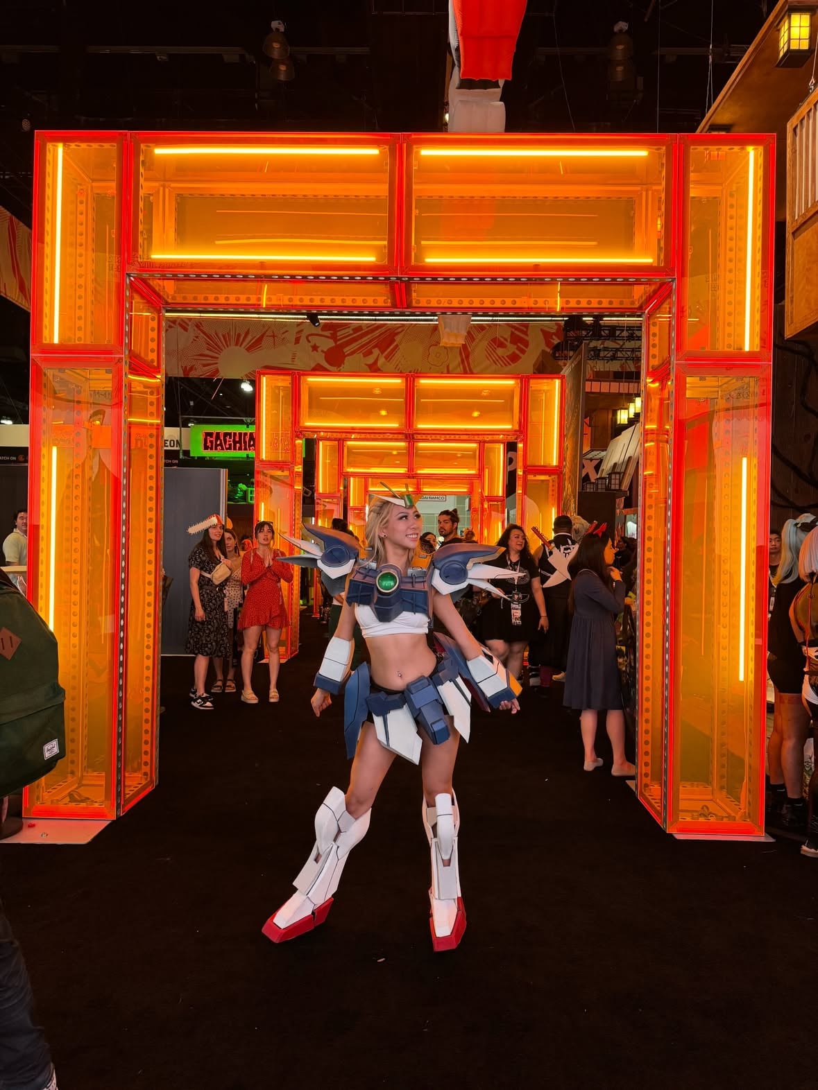 Full body anime cosplay portrait photographed indoors at Fanime convention during Memorial Day weekend. Blonde female cosplayer posing confidently in detailed blue and white mecha armor inspired by Gundam-style anime aesthetics. The cosplay features large futuristic shoulder armor pieces, layered waist armor, armored shin guards, gauntlets, and a gold anime crown headpiece with green gem detailing. The cosplayer is holding a long white sci-fi sword while standing in front of an urban graffiti-style convention backdrop with neon green spray paint, red graffiti art, and cyberpunk street-art textures. The armor appears handmade from EVA foam with detailed painted panels and clean geometric construction. The cosplay combines futuristic anime warrior styling with convention fashion and craftsmanship. Long curled blonde hair, glam makeup, dramatic lashes, and soft pink lip makeup create a polished convention look. White crop top styling underneath the armor gives the cosplay a modern anime heroine aesthetic. Lighting has cinematic convention photography vibes with cool blue tones and soft shadows. Background includes convention attendees walking through the expo hall, creating authentic anime convention atmosphere. Fanime cosplay photography, anime convention fashion, Gundam cosplay, mecha armor cosplay, anime girl cosplay, handcrafted EVA foam armor, cosplay craftsmanship, anime expo style portrait photography, convention floor photography, kawaii anime aesthetic, sci-fi cosplay design, gaming convention outfit inspiration, anime armor build, cosplay creator community, otaku culture, convention weekend vibes, cosplay influencer photography, Memorial Day weekend convention energy, fantasy armor fashion, cyberpunk anime styling, anime warrior princess aesthetic.