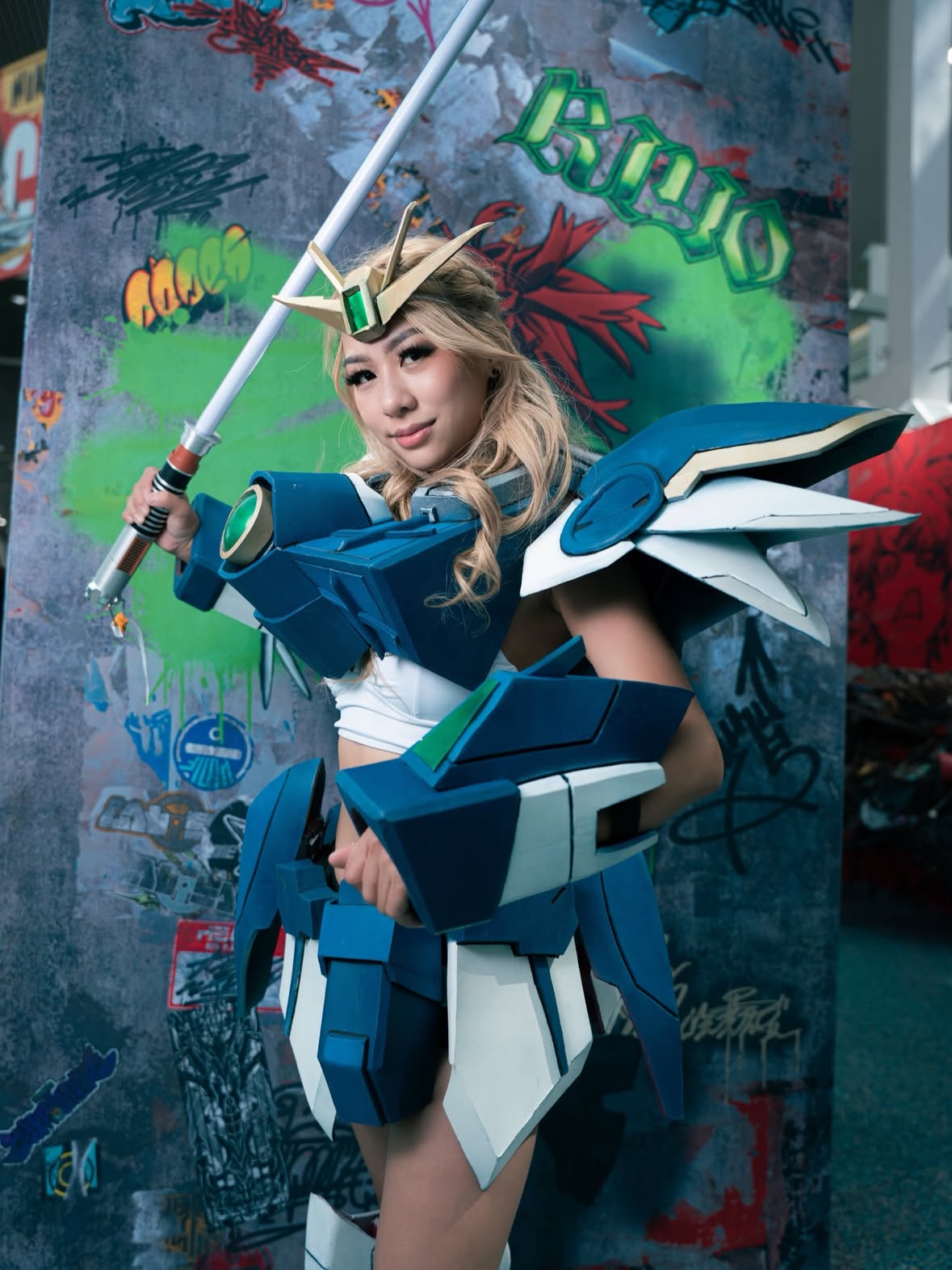 Vertical cosplay portrait of female anime cosplayer posing with oversized futuristic sword at Fanime anime convention. Blonde cosplayer wearing elaborate blue and white Gundam-inspired mecha armor with large shoulder wings, armored arm gauntlets, detailed waist plating, and futuristic anime battle accessories. Cosplay features handcrafted EVA foam armor with angular sci-fi panel details and metallic painted effects. The cosplayer is smiling confidently while holding a large anime-style weapon diagonally across the frame, creating dynamic action pose energy. Gold anime crown headpiece with green gemstone centerpiece adds fantasy anime queen aesthetic. Long curled blonde hair styled glam with convention-ready makeup including dramatic eyelashes and soft glam beauty look. Background features colorful graffiti wall with neon green spray paint, red anime-inspired graffiti art, and textured cyberpunk urban styling inside convention center hallway. Cinematic anime convention photography with cool teal and blue color grading gives the image high-energy cosplay photoshoot vibes. Fanime 2026 cosplay content, anime convention photography, mecha cosplay armor, Gundam cosplay inspiration, anime sword cosplay, female anime warrior aesthetic, handcrafted cosplay build, convention floor photoshoot, cosplay influencer style, otaku fashion culture, sci-fi anime cosplay, gaming expo outfit, anime armor details, fantasy anime character vibes, kawaii cosplay creator, cosplay craftsmanship community, anime event weekend, convention cosplay model photography, cyberpunk anime visuals, cosplay photography inspiration, viral anime convention content.